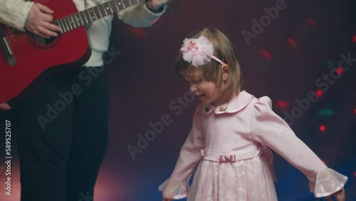 Little girl on stage in pink vintage dress, she dances, her father plays acoustic guitar. Color music is shining. Performance on stage. Early development of children