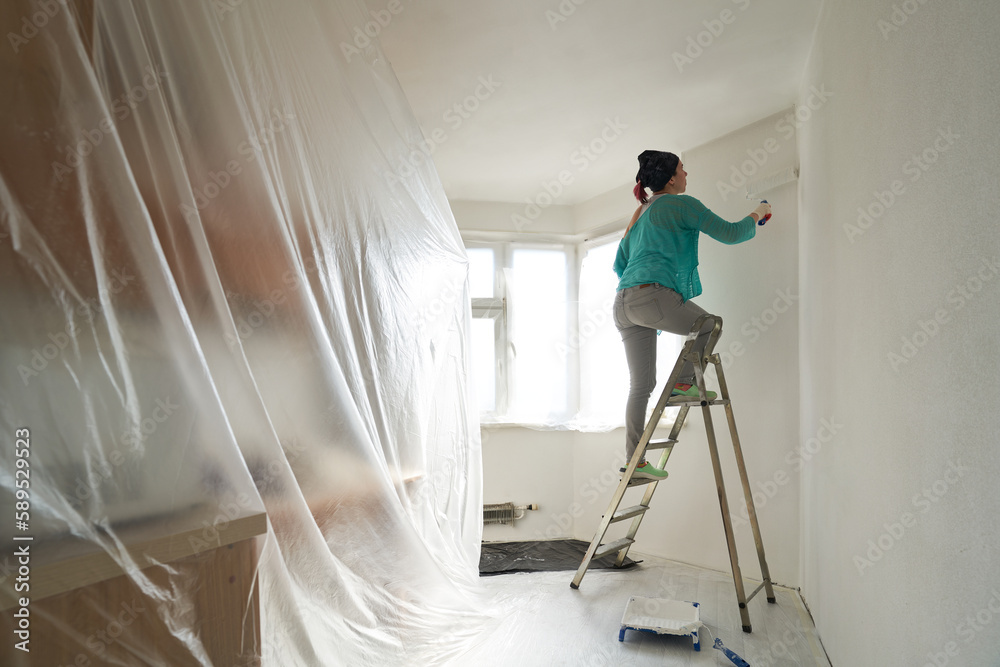Woman stands on the stairs and paints the wall with a roller - obrazy, fototapety, plakaty 