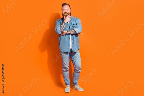 Full length photo of smart clever man wear jeans shirt arm chin smiling isolated orange color background