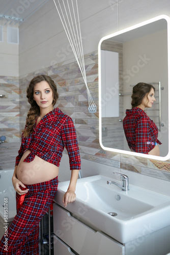 A pregnant woman in the bathroom. Female dressed in red pajama and open belly enjoying her pregnancy state
