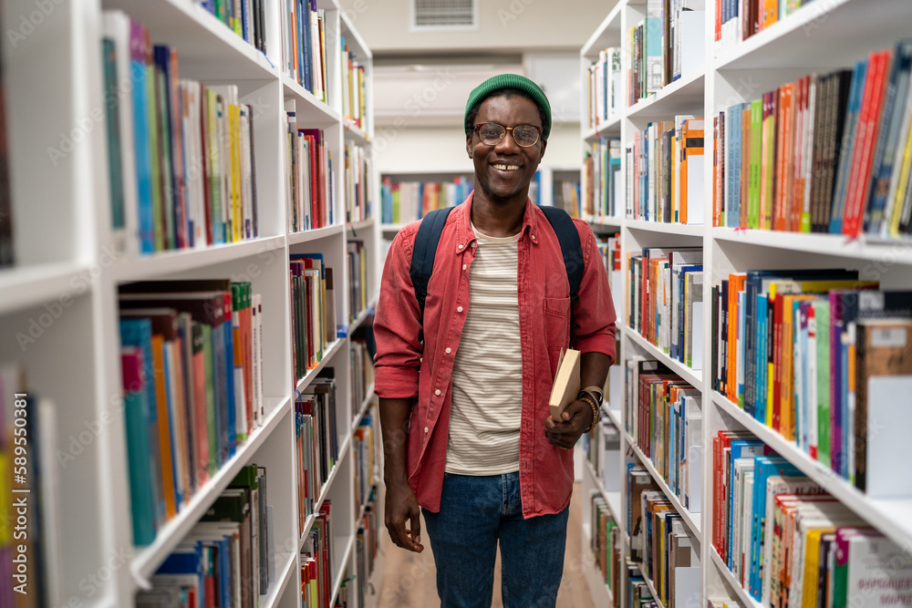 Smiling african american man student holding book in university library standing between bookshelves. Life in campus, education, self-development reading books studying learning researching concept. Smiling african american man student holding book in university library standing between bookshelves. Life in campus, education, self-development reading books studying learning researching concept.
