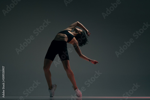 Flexible, athletic young woman with strong, relief, slim body in sportswear training again dark grey studio background. Concept of sportive lifestyle, beauty, body care, fitness, health