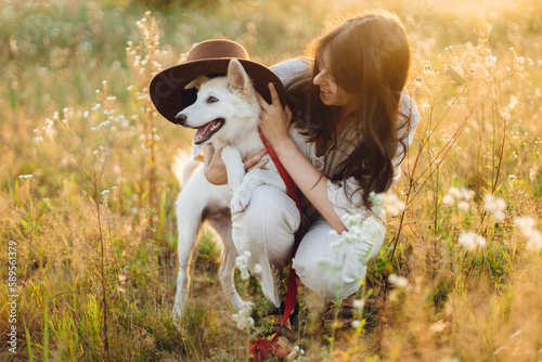Stylish happy woman playing with cute dog with hat among wildflowers in sunset light. Summer travel with pet. Young carefree female having fun with white danish spitz in summer meadow
