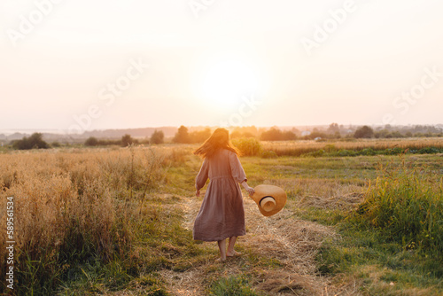 Stylish woman with straw hat dancing at oat field in sunset light. Atmospheric happy moment. Young female in rustic linen dress relaxing in evening summer countryside  rural life