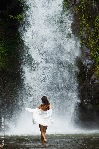 A young girl in a white shirt and swimsuit admires the waterfall, rear view.