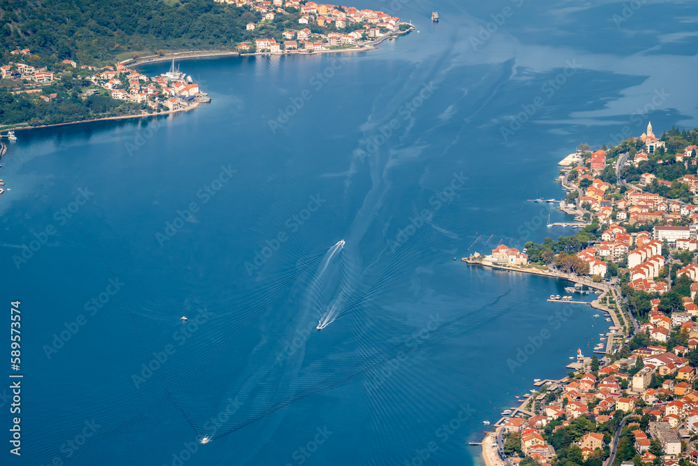 Amazing view from above of motor boats in the sky blue sea of the Bay of Kotor in Montenegro, with a picturesque coastline, red roofs of houses and the marina with boats.