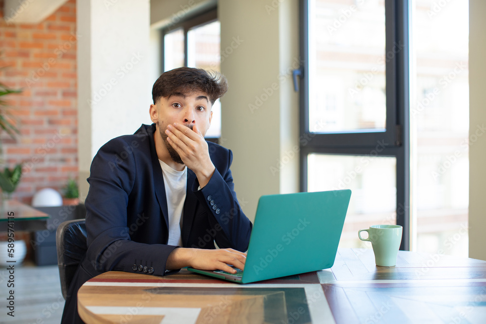 young handsome man covering mouth with a hand and shocked or surprised expression. freelance concept with laptop young handsome man covering mouth with a hand and shocked or surprised expression. freelance concept with laptop