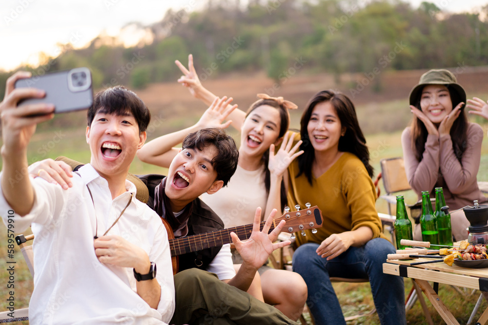 Obraz premium Group of happy Asian young men and women enjoy camping and party together, friends sitting at the park beside lake and enjoy grill a barbecue - stake and playing guitar together.