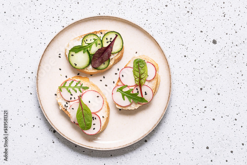 Bright juicy spring and summer snack. Set of bruschetta with cream cheese, cucumbers, small radish and fresh salad leaves on light stone background. Healthy homemade food ideas.