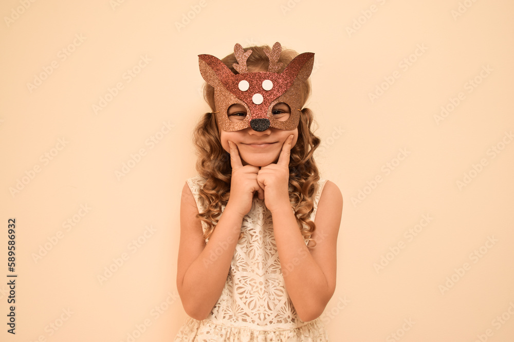 Little cute girl in a carnival masquerade deer mask made of shiny glitter foamiran on a beige background. Happy child in deer costume. The child is preparing for the masquerade. Little cute girl in a carnival masquerade deer mask made of shiny glitter foamiran on a beige background. Happy child in deer costume. The child is preparing for the masquerade.