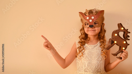 Fototapeta Naklejka Na Ścianę i Meble -  Little cute girl in a carnival masquerade deer mask made of shiny glitter foamiran on a beige background. Happy child in deer costume. The child is preparing for the masquerade.
