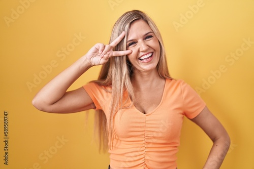 Young woman standing over yellow background doing peace symbol with fingers over face, smiling cheerful showing victory