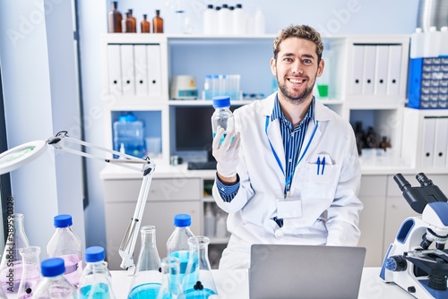Young man scientist using laptop holding bottle at laboratory