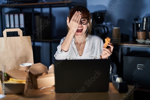 Young beautiful woman working using computer laptop and eating delivery food covering one eye with hand  confident smile on face and surprise emotion.