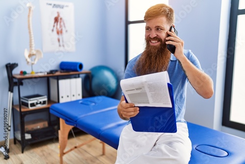 Young redhead man wearing physiotherapist uniform talking on the smartphone at physiotherapy clinic photo