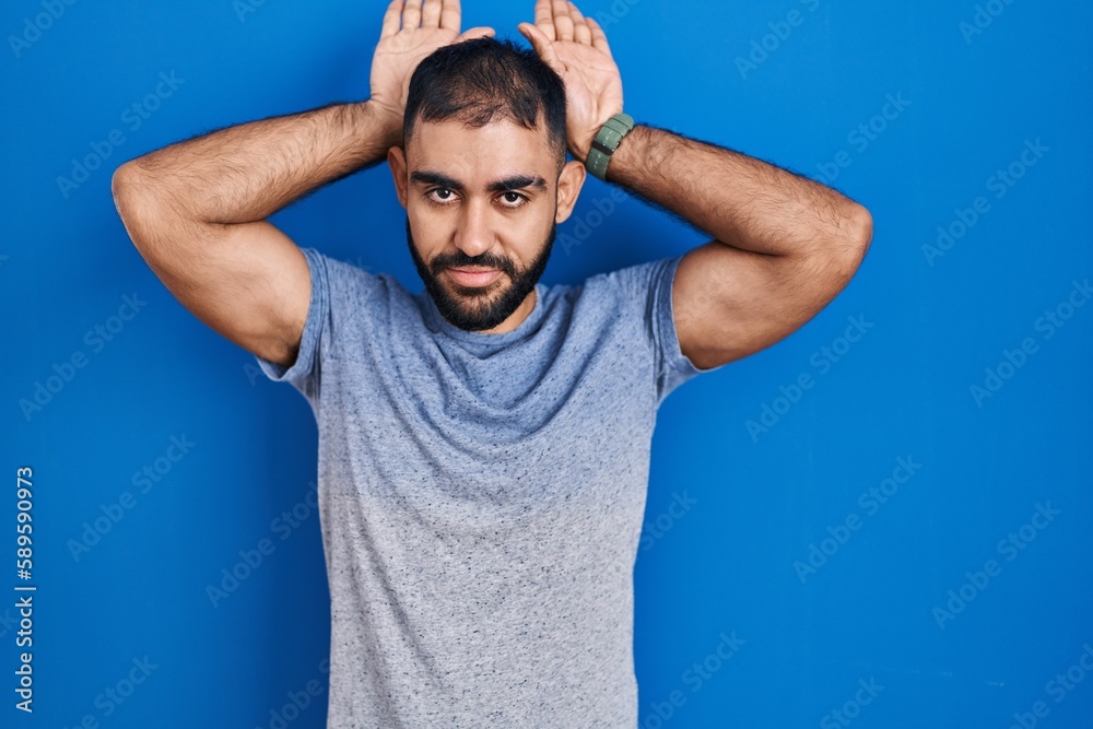 Middle east man with beard standing over blue background doing bunny ears gesture with hands palms looking cynical and skeptical. easter rabbit concept. Middle east man with beard standing over blue background doing bunny ears gesture with hands palms looking cynical and skeptical. easter rabbit concept.