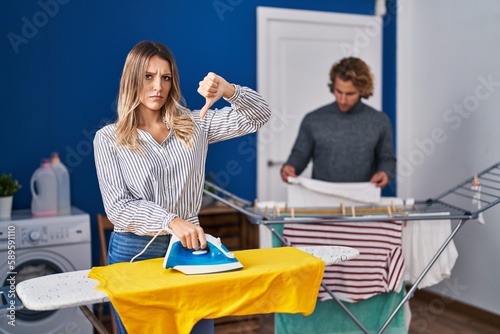 Couple ironing clothes at laundry room with angry face, negative sign showing dislike with thumbs down, rejection concept
