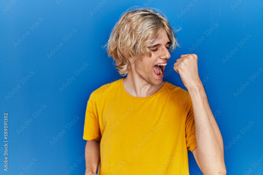 Young modern man standing over blue background very happy and excited doing winner gesture with arms raised, smiling and screaming for success. celebration concept. Young modern man standing over blue background very happy and excited doing winner gesture with arms raised, smiling and screaming for success. celebration concept.