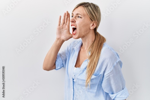 Beautiful blonde woman standing over white background shouting and screaming loud to side with hand on mouth. communication concept.