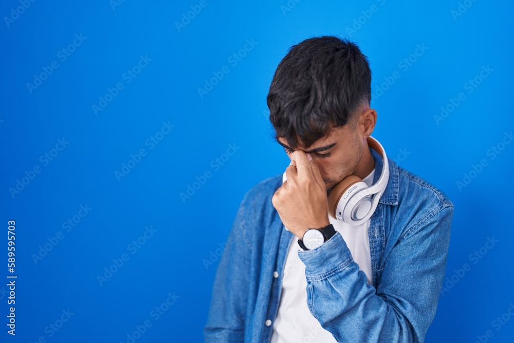 Young hispanic man standing over blue background tired rubbing nose and eyes feeling fatigue and headache. stress and frustration concept.