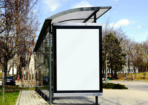 bus shelter at a busstop. blank billboard ad display. empty white lightbox sign. glass and aluminum frame structure. city transit station. urban street and green park setting. outdoor advertising.