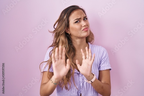 Young hispanic woman standing over pink background moving away hands palms showing refusal and denial with afraid and disgusting expression. stop and forbidden.