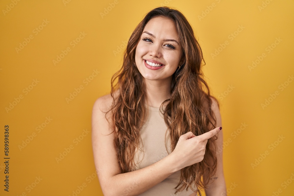Young hispanic woman standing over yellow background cheerful with a smile of face pointing with hand and finger up to the side with happy and natural expression on face Young hispanic woman standing over yellow background cheerful with a smile of face pointing with hand and finger up to the side with happy and natural expression on face