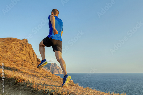 back male runner run uphill mountain trail in blue sky background, summer jogging along coast at sunset