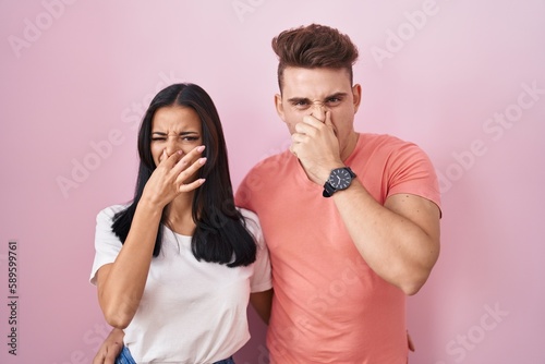 Young hispanic couple standing over pink background smelling something stinky and disgusting, intolerable smell, holding breath with fingers on nose. bad smell