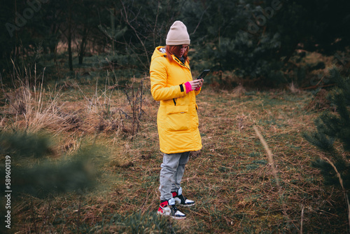 A girl, a woman in a yellow winter long jacket stands in the middle of wild nature, forest. He looks at his smartphone. Concept of navigation, communication. Get lost.