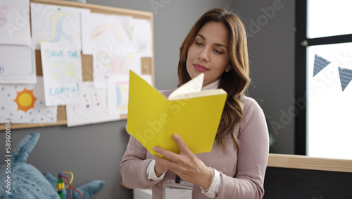 Young beautiful hispanic woman preschool teacher reading book standing at kindergarten