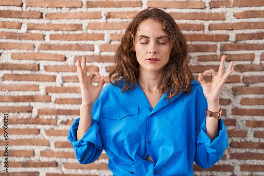 Beautiful brunette woman standing over bricks wall relaxed and smiling with eyes closed doing meditation gesture with fingers. yoga concept. Beautiful brunette woman standing over bricks wall relaxed and smiling with eyes closed doing meditation gesture with fingers. yoga concept.