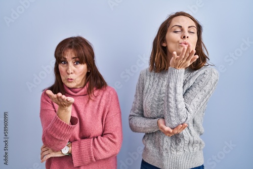 Mother and daughter standing over blue background looking at the camera blowing a kiss with hand on air being lovely and sexy. love expression.