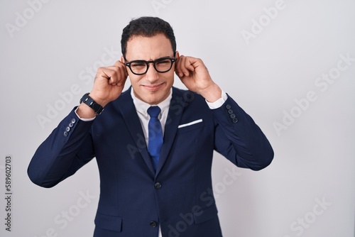 Young hispanic man wearing suit and tie covering ears with fingers with annoyed expression for the noise of loud music. deaf concept.