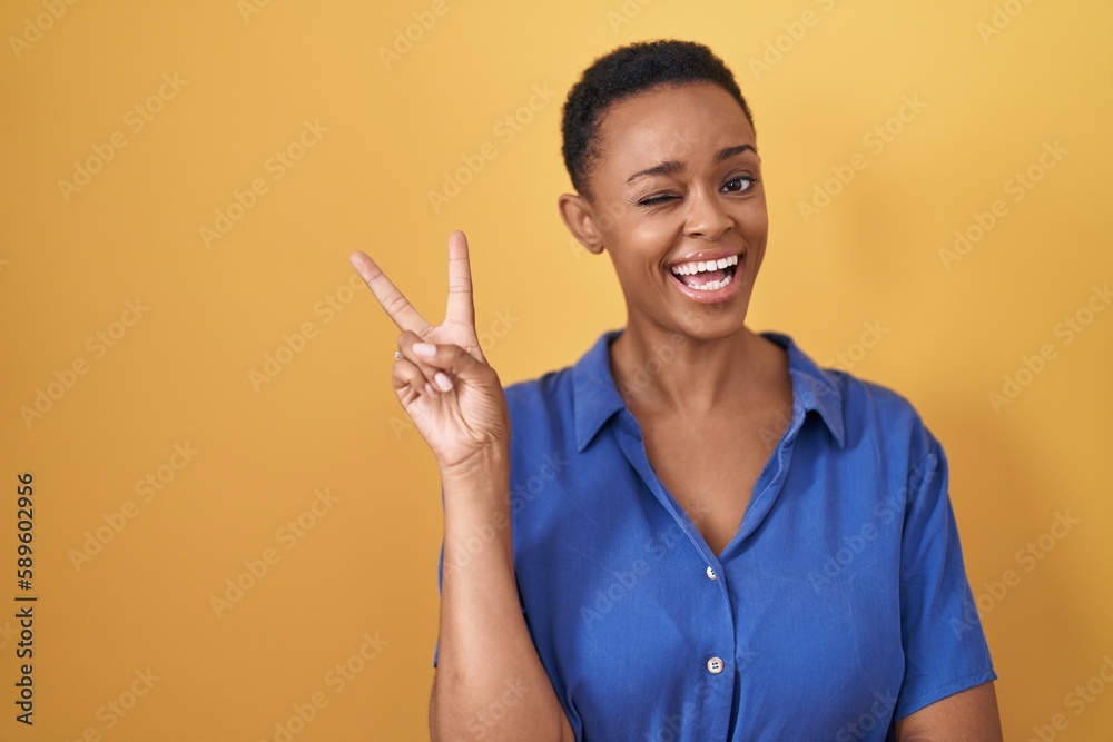 African american woman standing over yellow background smiling with happy face winking at the camera doing victory sign with fingers. number two. African american woman standing over yellow background smiling with happy face winking at the camera doing victory sign with fingers. number two.