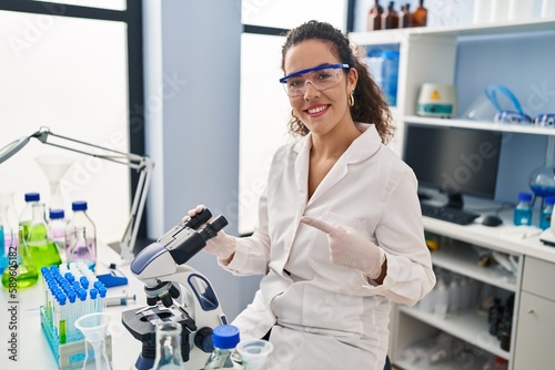 Young hispanic woman working at scientist laboratory smiling happy pointing with hand and finger