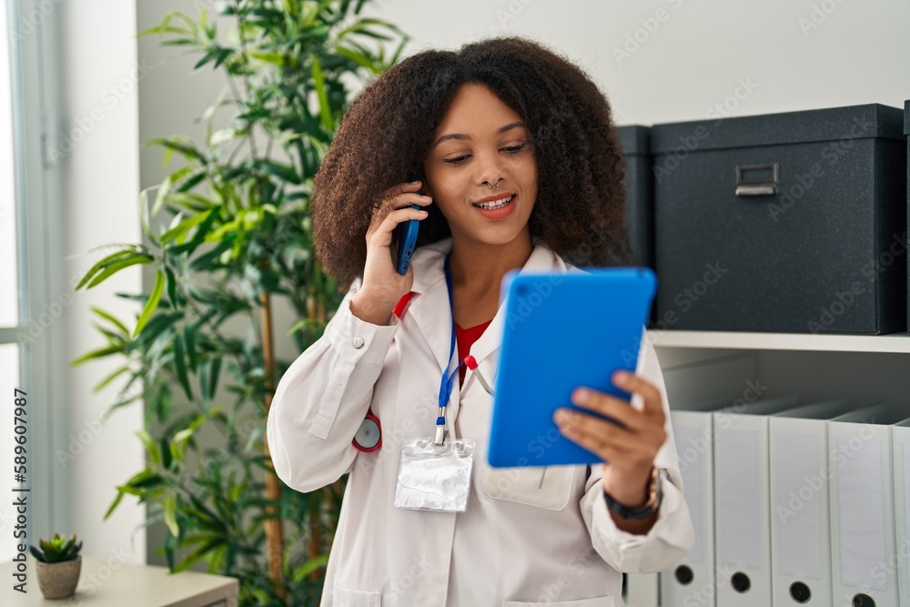 Young african american woman wearing doctor uniform talking on the smartphone using touchpad at clinic