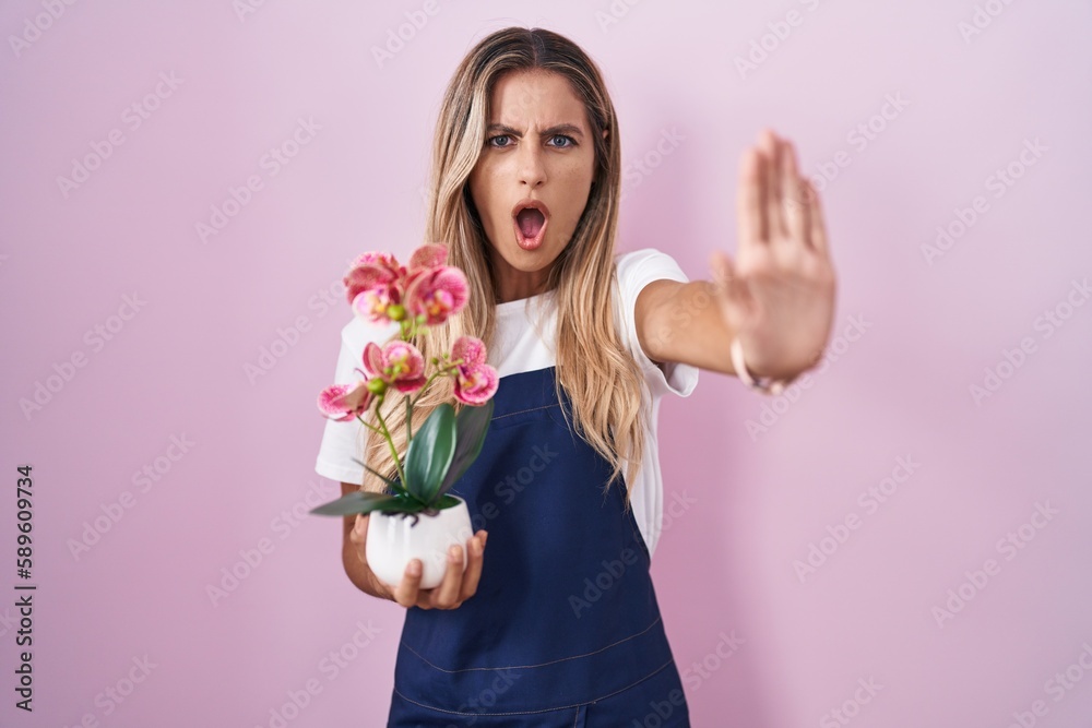 Young blonde woman wearing gardener apron holding plant doing stop gesture with hands palms, angry and frustration expression Young blonde woman wearing gardener apron holding plant doing stop gesture with hands palms, angry and frustration expression
