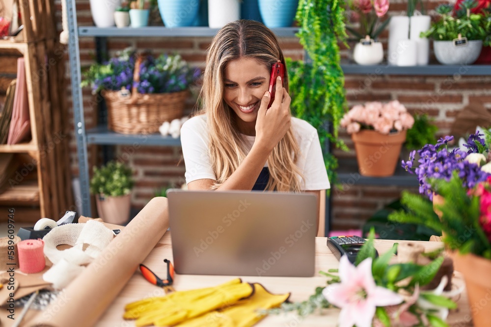 Young blonde woman florist talking on smartphone using laptop at florist