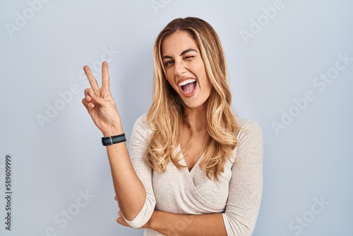 Foto Young blonde woman standing over isolated background smiling with happy face winking at the camera doing victory sign with fingers