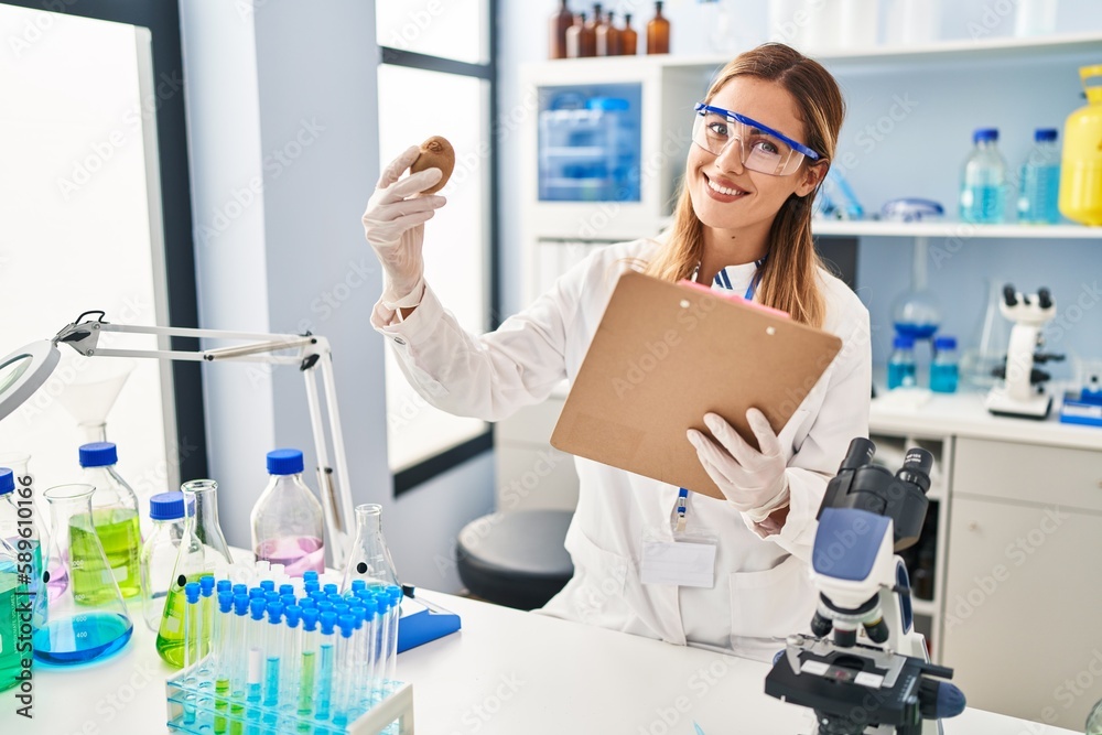Young blonde woman wearing scientist uniform holding kiwi and clipboard at laboratory