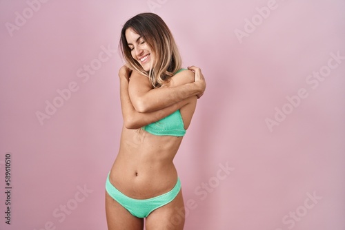 Young hispanic woman wearing bikini over pink background hugging oneself happy and positive, smiling confident. self love and self care