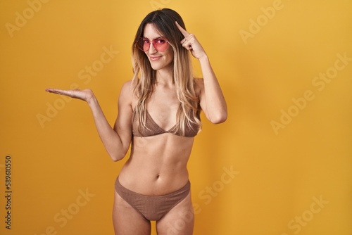 Young hispanic woman wearing bikini over yellow background confused and annoyed with open palm showing copy space and pointing finger to forehead. think about it.