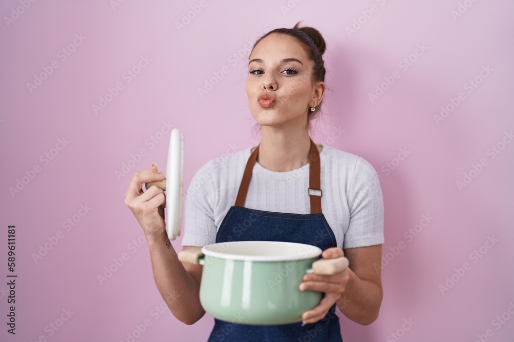 Young hispanic girl wearing apron holding cooking pot looking at the camera blowing a kiss being lovely and sexy. love expression. Young hispanic girl wearing apron holding cooking pot looking at the camera blowing a kiss being lovely and sexy. love expression.