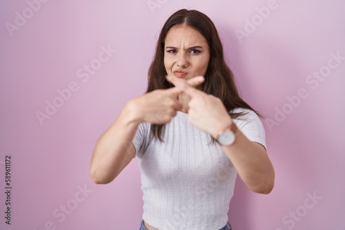 Young hispanic girl standing over pink background rejection expression crossing fingers doing negative sign