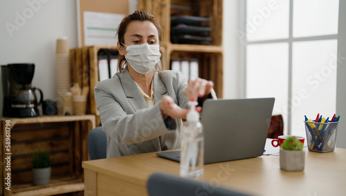 Middle age hispanic woman business worker wearing medical mask using sanitizer gel hands at office