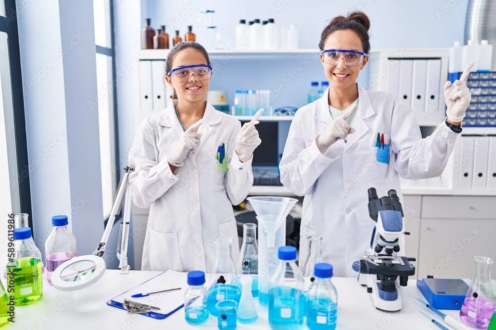 Young mother and daughter at scientist laboratory smiling and looking at the camera pointing with two hands and fingers to the side. Young mother and daughter at scientist laboratory smiling and looking at the camera pointing with two hands and fingers to the side.