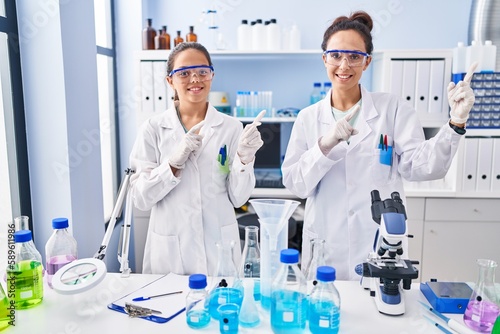 Young mother and daughter at scientist laboratory smiling and looking at the camera pointing with two hands and fingers to the side.