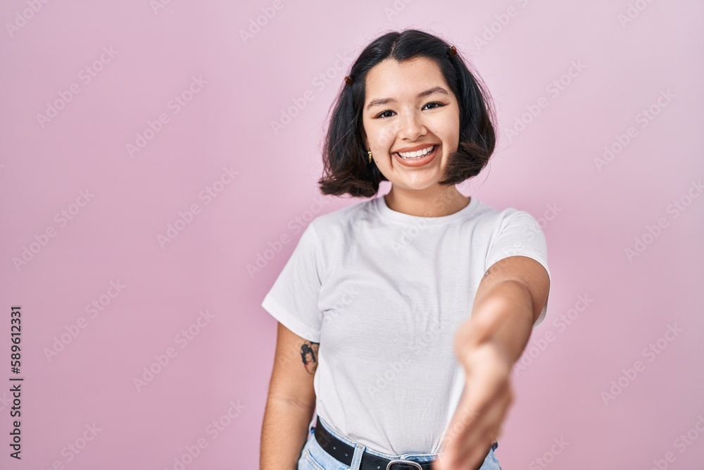 Young hispanic woman wearing casual white t shirt over pink background smiling friendly offering handshake as greeting and welcoming. successful business.
