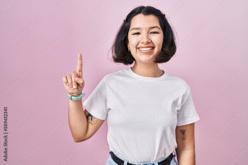 Young hispanic woman wearing casual white t shirt over pink background showing and pointing up with finger number one while smiling confident and happy. Young hispanic woman wearing casual white t shirt over pink background showing and pointing up with finger number one while smiling confident and happy.